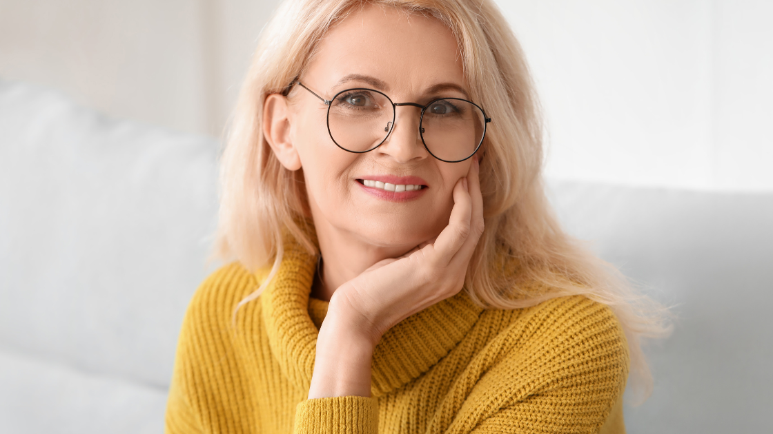 Frau mit blonden Haaren und einer Brille trägt einen gelben Pullover und schaut in die Kamera.